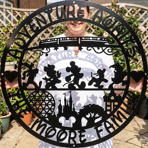 Puede incluir: Un letrero de pared circular de metal negro con el texto ADVENTURE AWAITS en la parte superior y THE MOORE FAMILY en la parte inferior. El diseño incluye siluetas de personajes de dibujos animados, un monorraíl, un castillo, un domo y un árbol.