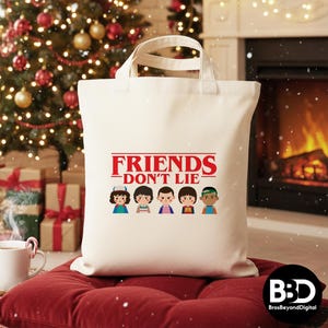 May include: Cream-colored tote bag with the text "FRIENDS DON'T LIE" in red, with cartoon character illustrations below. The bag has white handles and is sitting on a red cushion. A Christmas tree and fireplace are in the background.