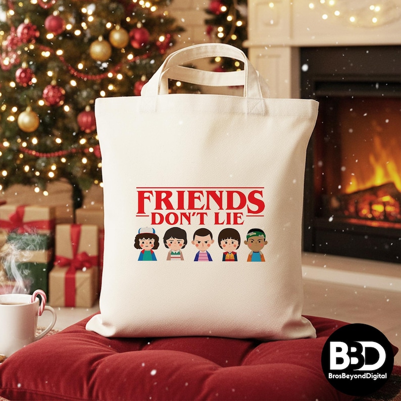 May include: Cream-colored tote bag with the text "FRIENDS DON'T LIE" in red, with cartoon character illustrations below. The bag has white handles and is sitting on a red cushion. A Christmas tree and fireplace are in the background.