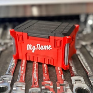 May include: A red and black tool organizer with the text "My Name" in white, resting on a set of wrenches. The organizer is rectangular with a textured top and is designed to hold tools. The wrenches are silver and red.