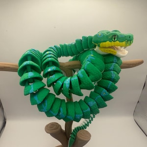 May include: A green and blue articulated snake toy coiled around a brown branch. The snake's head is yellow and green with an open mouth. The toy is made of multiple connected segments, allowing for flexible posing.