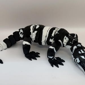 May include: A black and white plush lizard toy. The toy features a long tail and four clawed feet. The lizard's body is mainly black with white markings, and the tail is striped in black and white. It is positioned on a white surface.