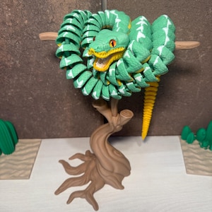 May include: A green and white articulated snake toy with an open yellow mouth and orange eyes. The snake is coiled around a wooden branch, attached to a brown tree base. The snake's tail is yellow.