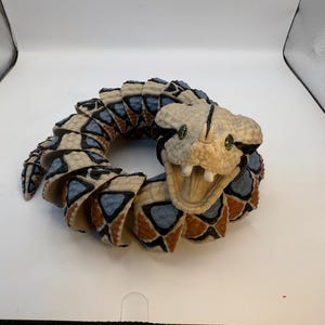 May include: A coiled snake ornament with a detailed design. The snake's body is segmented with a pattern of beige, blue, and brown shapes. The head is beige with green eyes and an open mouth, revealing teeth. The ornament is a decorative item.