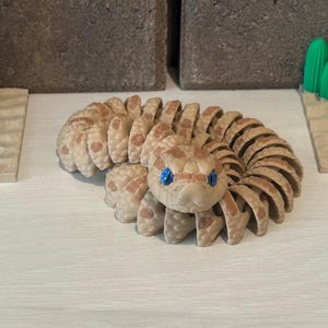 May include: A coiled, tan snake figurine with a detailed segmented body and blue eyes. The snake is on a white surface, with miniature desert-themed decorations in the background.