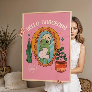Könnte beinhalten: Ein gerahmter rosa Kunstdruck mit einem Cartoon-Frosch im Handtuch, der in einen Spiegel schaut. Der Druck enthält den Text "HELLO GORGEOUS" und "YOU LOOK SO CUTE". Ein grünes Handtuch hängt an einem Haken, und eine Topfpflanze befindet sich rechts.