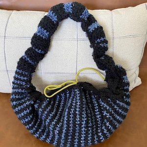 May include: A handmade, black and blue crocheted bag with a rounded shape and a yellow drawstring closure. The bag features vertical stripes of black and blue yarn, and a looped handle. The bag is resting on a white pillow with a plaid pattern.