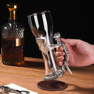 May include: A clear glass beer mug with a silver dragon handle and wing accents. The dragon's tail wraps around the base of the mug. A bottle with a gold stopper is in the background.