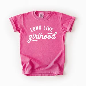 May include: A bright pink t-shirt with the words "LONG LIVE girlhood" in white script. The shirt has a crew neck and short sleeves with rolled cuffs. The fabric appears soft and slightly faded.