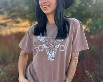 May include: A brown t-shirt with a white bull skull graphic and the word "HOWDY" printed on it. The person is wearing a black hat and blue jeans. The background is a blurred outdoor scene.