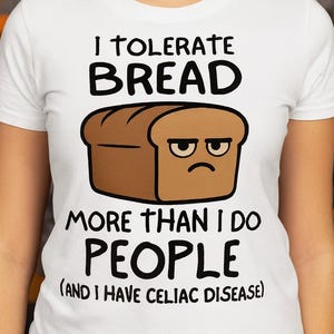 May include: White t-shirt with a humorous graphic. The design features a loaf of bread with a grumpy face and the text "I TOLERATE BREAD MORE THAN I DO PEOPLE (AND I HAVE CELIAC DISEASE)".