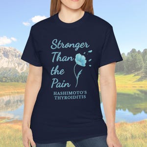 May include: Navy blue t-shirt with the text "Stronger Than the Pain" and a light blue flower graphic. The text "Hashimoto's Thyroiditis" is below the main text. The shirt is a crew neck style.
