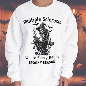Puede incluir: Sudadera blanca con un diseño de Halloween. El diseño presenta una bruja esqueleto con un caldero, murciélagos y el texto "Multiple Sclerosis Where Every Day is Spooky Season."