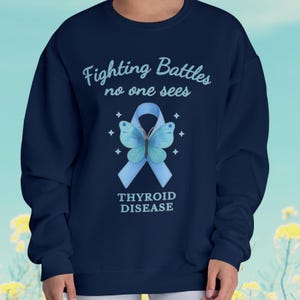 Op de afbeelding: Marineblauw sweatshirt met de tekst "Fighting Battles no one sees" boven een blauw lint en vlinderontwerp. De woorden "Thyroid Disease" staan eronder. De sweatshirt is effen van kleur.