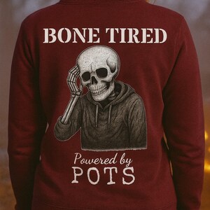 Könnte beinhalten: Burgunderfarbener Kapuzenpullover mit einer Illustration eines Skeletts. Die Worte "BONE TIRED" stehen über dem Skelett und "Powered by POTS" darunter. Das Skelett ist mit der Hand am Kopf dargestellt.