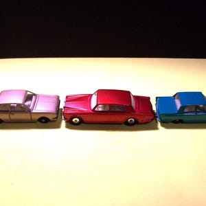 May include: Three die-cast toy cars arranged in a horizontal row. The car on the left is metallic light purple, the center car is metallic red, and the car on the right is metallic blue. Each vehicle features four black wheels and white windows.