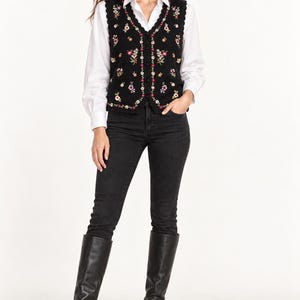 May include: A black sleeveless vest featuring a V-neckline with a scalloped edge and a button-down front. The vest is decorated with an embroidered floral pattern in shades of pink, yellow, white, and green across the front panels.