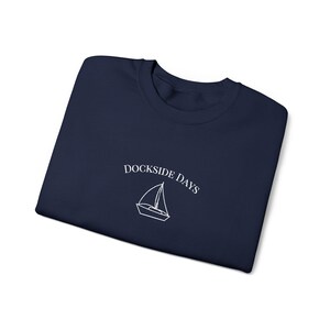 May include: Navy blue crew neck sweatshirt with the text "Dockside Days" above a white sailboat graphic. The sweatshirt is folded and displayed on a white background.