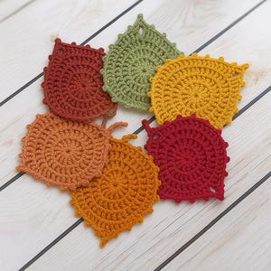 May include: A collection of six crocheted leaf-shaped coasters in autumnal colors: rust, olive green, golden yellow, orange, and red. Each coaster features a textured, circular design with a leaf-like shape and a small loop for hanging.