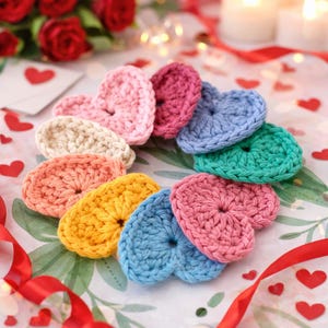 Peut inclure: Un ensemble de cœurs crochetés à la main de différentes couleurs, dont rose, bleu, jaune et vert. Les cœurs sont disposés en cercle sur un fond floral, avec un ruban rouge et des confettis en forme de cœur.