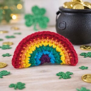 Pode incluir: Um arco-íris de crochê em vermelho, laranja, amarelo, verde, azul e roxo. O arco-íris é o ponto focal, rodeado por moedas de ouro, trevos verdes e um pote preto cheio de moedas de ouro, evocando o tema do Dia de São Patrício.