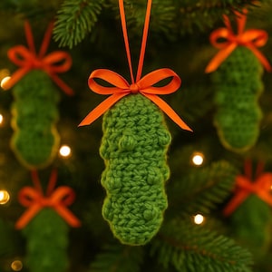 Puede incluir: Adornos navideños verdes tejidos a ganchillo con forma de pepinillos, cada uno con un lazo de cinta naranja para colgar. Los adornos cuelgan de un árbol de Navidad, con luces suaves y borrosas en el fondo.
