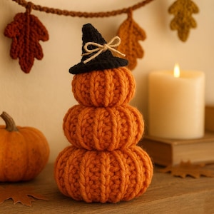Puede incluir: Una pila de tres calabazas naranjas tejidas, coronadas con un sombrero de bruja negro con un lazo marrón. A la izquierda hay una pequeña calabaza naranja. Una vela blanca encendida y una guirnalda de hojas otoñales en el fondo. Decoración otoñal.