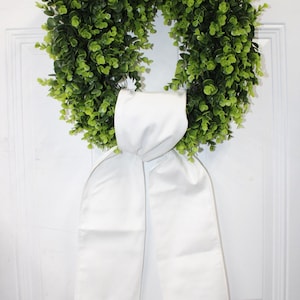 May include: A vibrant green wreath made of leafy foliage, accented with a large white satin bow. The wreath is circular and appears to be hanging on a white door, creating a fresh and welcoming aesthetic.