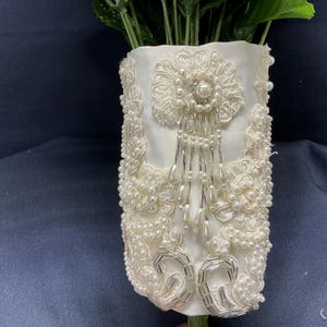 Può includere: Fodera per bouquet in tessuto avorio, riccamente impreziosita da perle e dettagli argentati. Il design presenta un motivo floreale centrale con una perla al centro, fili di perline a cascata e ricami intricati.