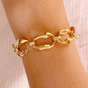 Könnte beinhalten: Ein goldfarbenes Armband mit einer Reihe von miteinander verbundenen, unregelmäßig ovalen Gliedern mit einer polierten, reflektierenden Oberfläche. Das Ketten-Design besteht aus sich wiederholenden, leicht gedrehten Metallschleifen.
