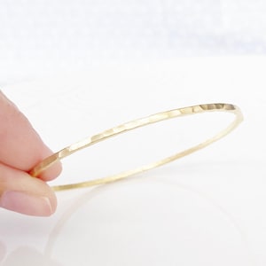 Pode incluir: Uma pulseira de bracelete dourada com textura martelada. A pulseira é fina e circular, com uma superfície ligeiramente irregular. O fundo é branco e uma mão segura a pulseira.