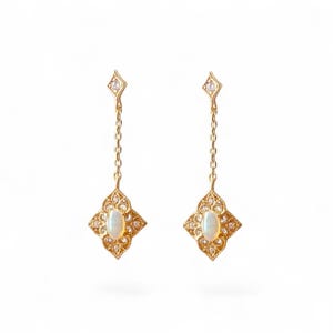 Könnte beinhalten: Ein Paar goldfarbene Ohrhänger. Jeder Ohrring hat eine rautenförmige Oberseite mit einem kleinen, klaren Stein, eine zarte Kette und ein filigranes Design mit einem zentralen ovalen Opalstein. Die Ohrringe sind vor weißem Hintergrund.