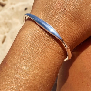 Könnte beinhalten: Ein polierter Armreif in Silberoptik mit einem glatten, geschwungenen und sich verjüngenden Design. Die Metalloberfläche reflektiert das Licht und zeigt ein elegantes, durchgehendes Band, das das Handgelenk mit einer offenen Struktur umschließt.
