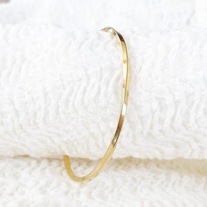 Op de afbeelding: Een delicate goudkleurige armband. De dunne, gehamerde metalen armband heeft een eenvoudig, minimalistisch ontwerp. De armband wordt gepresenteerd op een wit, getextureerd oppervlak.