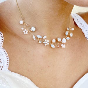 Op de afbeelding: Een goudkleurige ketting met een delicaat draadontwerp in de vorm van een rank, versierd met kleine witte bloemaccenten, heldere gefacetteerde kristallen en gladde ronde kralen. Het sieraad heeft een dunne gouden ketting en een flexibele draadbasis.