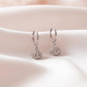 Può includere: Un paio di orecchini a cerchio color argento con pendenti a forma di conchiglia. Ogni pendente è incastonato con piccole pietre chiare e scintillanti che coprono la superficie della conchiglia, creando un aspetto strutturato e riflettente.
