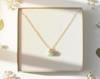 May include: A gold chain necklace featuring a small, faceted, pale green gemstone pendant. The necklace is displayed inside a square, cream-colored jewelry box with a soft, padded interior lining, presenting a delicate and refined appearance.