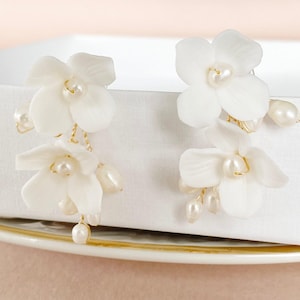 Könnte beinhalten: Ein Paar weiße Blumenohrringe mit Perlenakzenten. Jeder Ohrring hat mehrere weiße Blütenblätter und baumelnde Perlen, die durch goldfarbenen Draht verbunden sind. Die Ohrringe sind als Statement-Schmuck konzipiert.