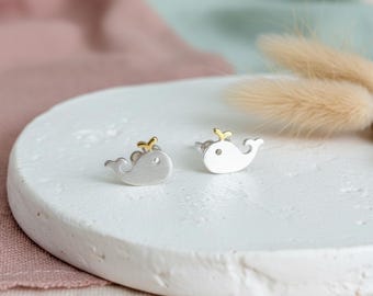 May include: A pair of silver-toned stud earrings shaped like whales, each featuring a small gold-toned accent on the top of the head. The earrings have a smooth, matte finish and are designed with a simple, rounded silhouette representing the whale body.