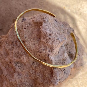 Peut inclure: Un bracelet manchette doré avec une texture martelée. Le bracelet ouvert repose sur une pierre brune rugueuse. Le bracelet a un design simple et élégant, avec une surface légèrement irrégulière.