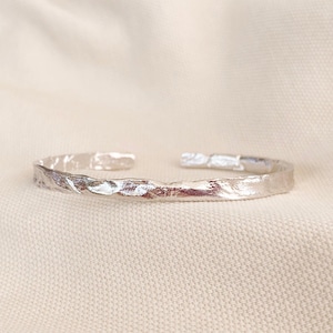 Pode incluir: Uma pulseira de punho prateada com uma aparência texturizada e martelada. O design aberto apresenta uma superfície ligeiramente irregular, que reflete a luz. A pulseira é exibida em um fundo neutro, branco sujo, destacando seu brilho metálico.