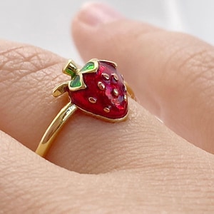 Könnte beinhalten: Ein goldfarbener Ring mit einem roten Erdbeer-Design mit grünen Blättern. Die Erdbeere hat eine strukturierte Oberfläche und ist auf einem dünnen, goldenen Band angebracht. Der Ring wird an einem Finger gezeigt.