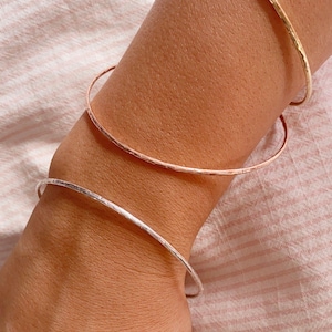 Könnte beinhalten: Nahaufnahme von drei dünnen, gehämmerten Metallarmreifen in Gold, Roségold und Silber. Die Armreifen werden an einem Handgelenk vor einem rosa-weiß gestreiften Hintergrund getragen. Die Armreifen haben eine strukturierte Oberfläche.