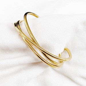 May include: A gold-colored cuff bracelet with a modern design. The bracelet features three curved, intersecting bands that create an open, geometric shape. The jewelry is displayed on a white, textured fabric.