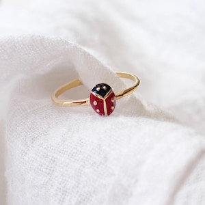 Op de afbeelding: Een goudkleurige ring met een lieveheersbeestje-ontwerp. Het lieveheersbeestje is rood met zwarte stippen en een zwarte kop. De ring staat tegen een witte, getextureerde achtergrond.