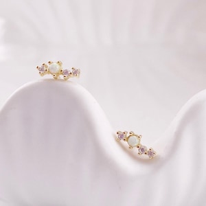 May include: Two gold-colored stud earrings, each featuring a central white opal stone flanked by three smaller pink stones. The earrings are displayed on a white, curved surface, creating a delicate and elegant aesthetic.