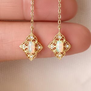 Può includere: Un paio di orecchini pendenti in oro con un cabochon di opale ovale al centro di una montatura in filigrana. Ogni orecchino è sospeso a una delicata catenina d'oro ed è impreziosito da piccole pietre rotonde trasparenti incastonate nel metallo.