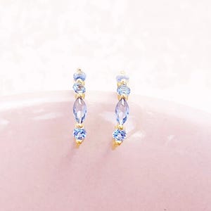 Puede incluir: Un par de pendientes dorados adornados con piedras preciosas azul claro. El diseño incorpora piedras redondas y en forma de marquesa, creando un aspecto elegante. Los pendientes se muestran sobre un fondo rosa suave.
