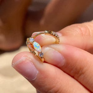 Op de afbeelding: Gouden oorringen met opaal- en kristalaccenten. Eén oorring heeft een marquise-vormige opaal en twee kleine kristallen. De andere oorring heeft een rij kleine kristallen. De oorbellen worden in een hand gehouden.