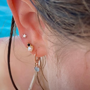 Könnte beinhalten: Nahaufnahme eines Ohrs mit mehreren Ohrringen. Oben befindet sich ein kleiner goldfarbener Sternstecker, gefolgt von einem Stecker mit einer Perle und einem schwarzen Stein. Ein goldener Creolen-Ohrring mit einem blauen Edelstein hängt darunter.